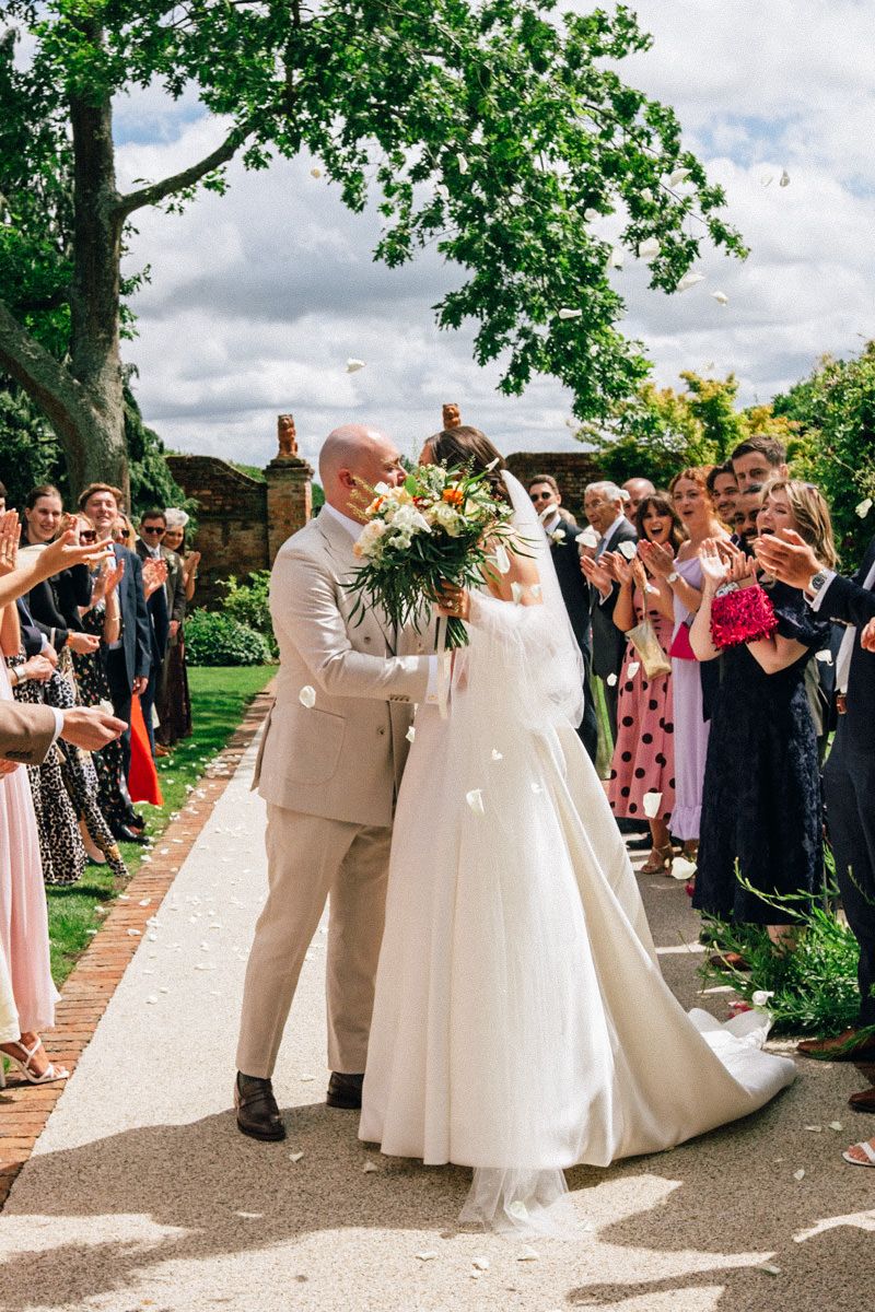 Newlyweds sharing first kiss during confetti exit after outdoor wedding ceremony.