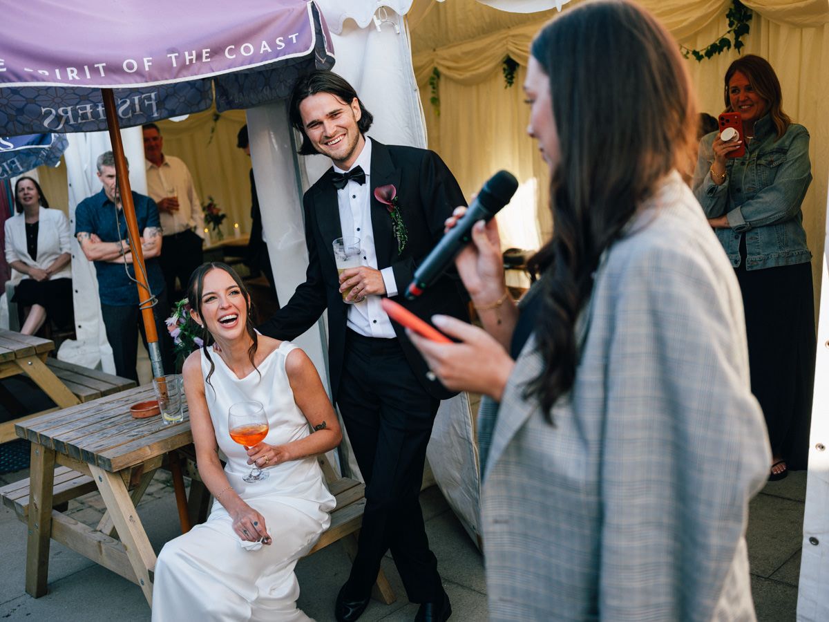 Bride laughing joyfully while seated as groom stands beside her during a wedding reception speech.