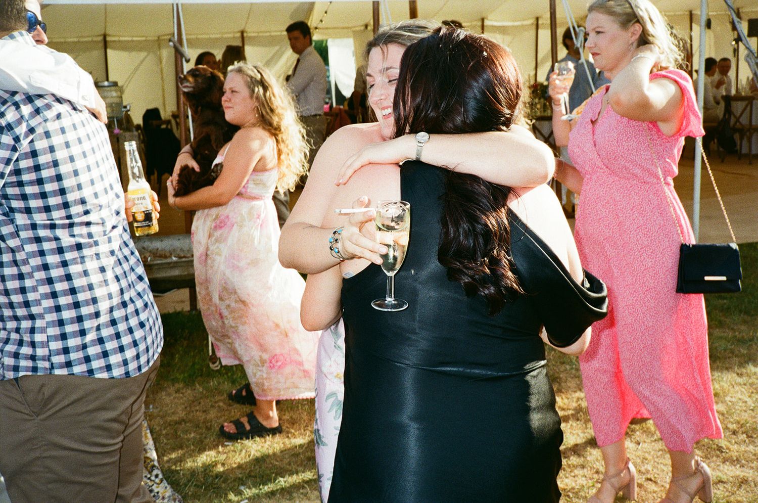 Wedding guests hugging and dancing inside marquee tent, woman holding wine glass in floral dress.