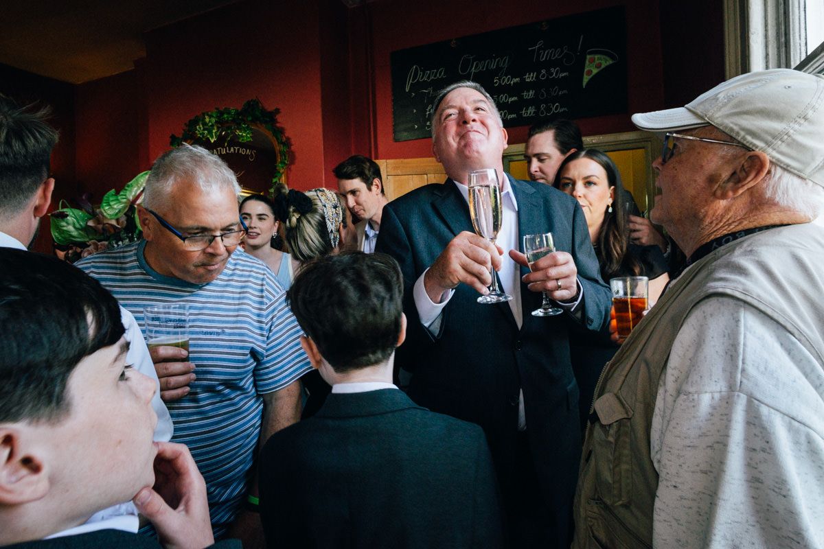 Wedding guests in formal attire laugh and toast with champagne glasses at an indoor reception.