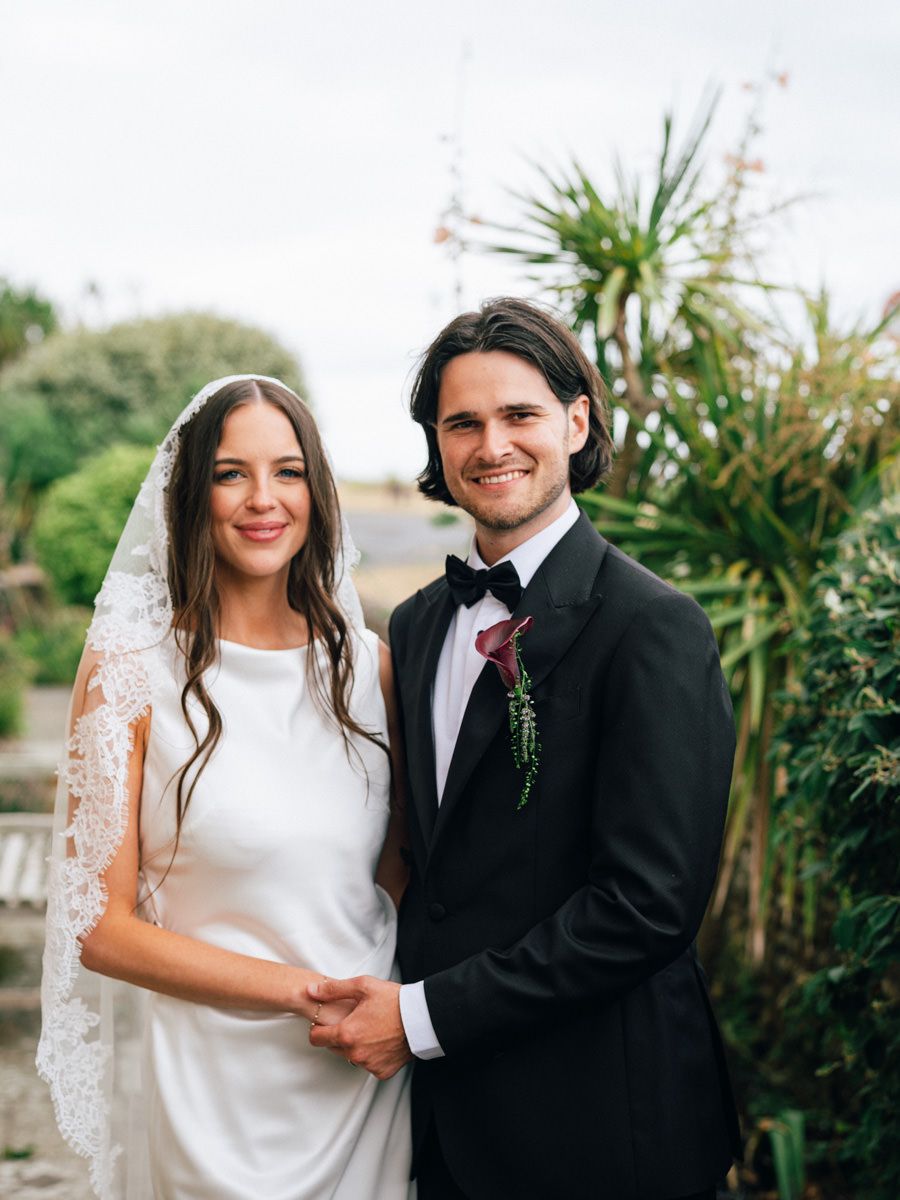 Smiling bride and groom posing together outdoors, bride wearing a lace mantilla veil.