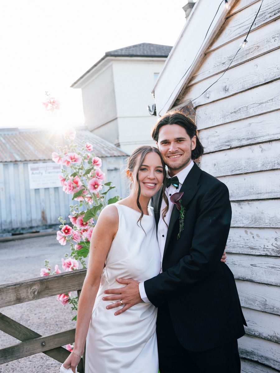Bride and groom embrace outdoors at golden hour beside a white wooden barn with pink flowers.