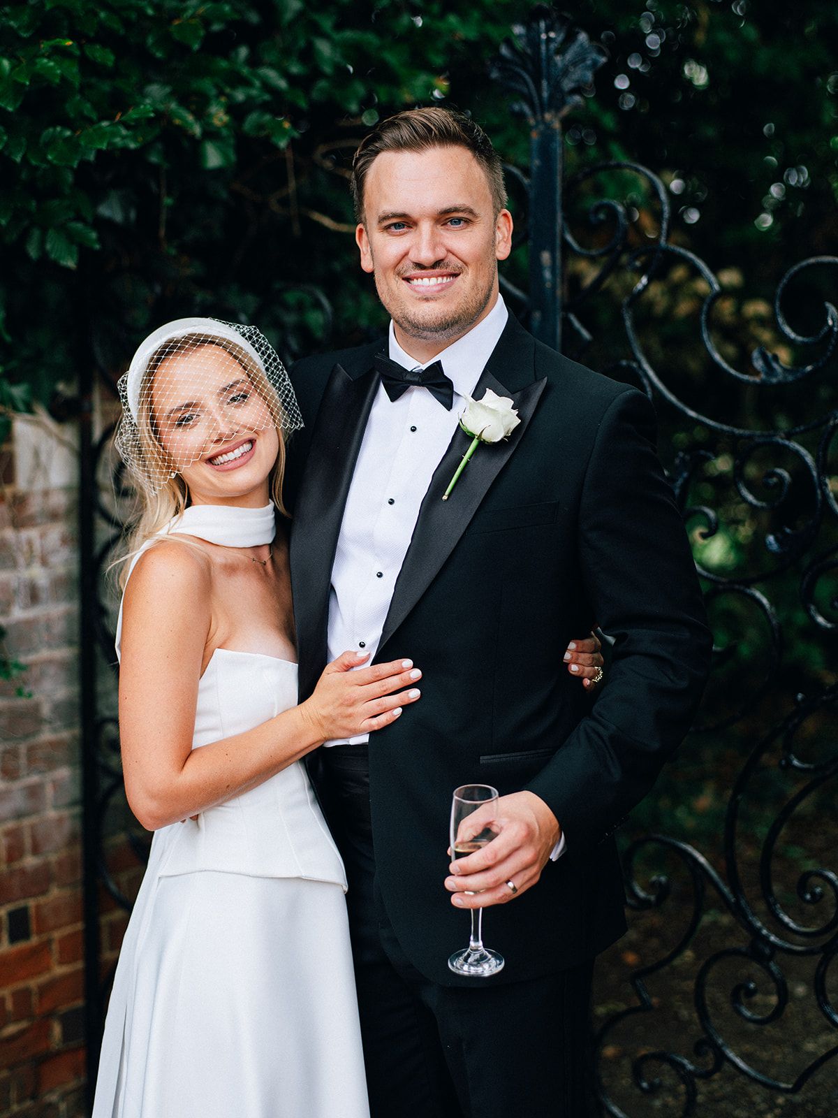 Elegant bride in white gown with birdcage veil and groom in black tuxedo posing together at an outdoor wedding venue.