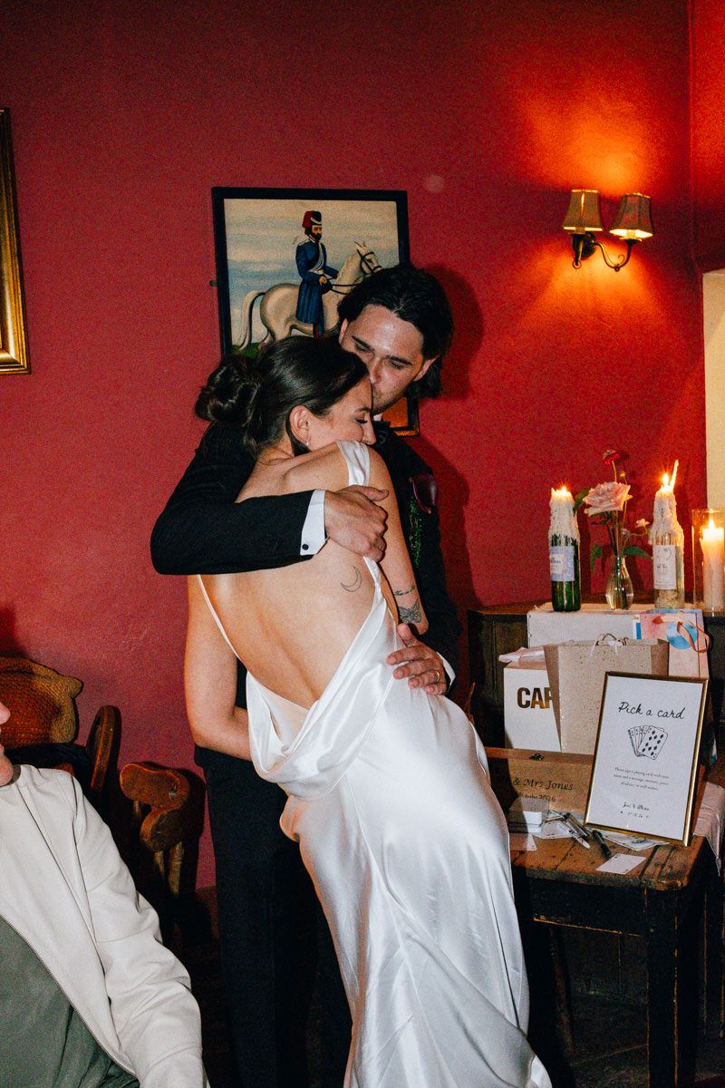 A groom embraces his bride in a backless white satin dress at their wedding reception.