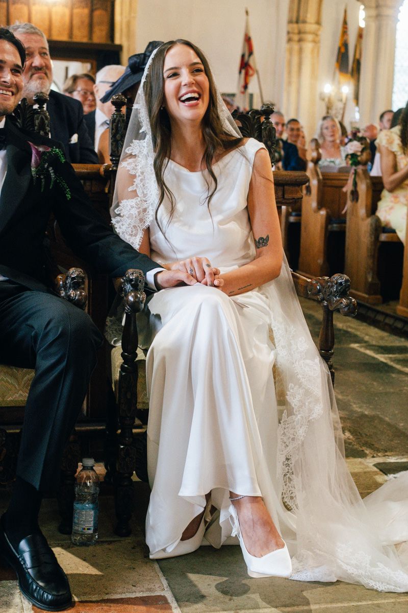 Laughing bride in white satin gown and lace veil seated during church wedding ceremony.