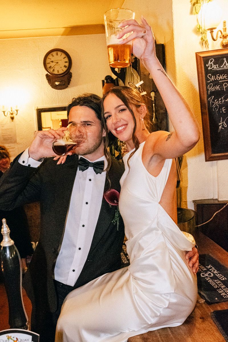 A bride and groom celebrate at a pub bar, raising glasses of beer on their wedding day.