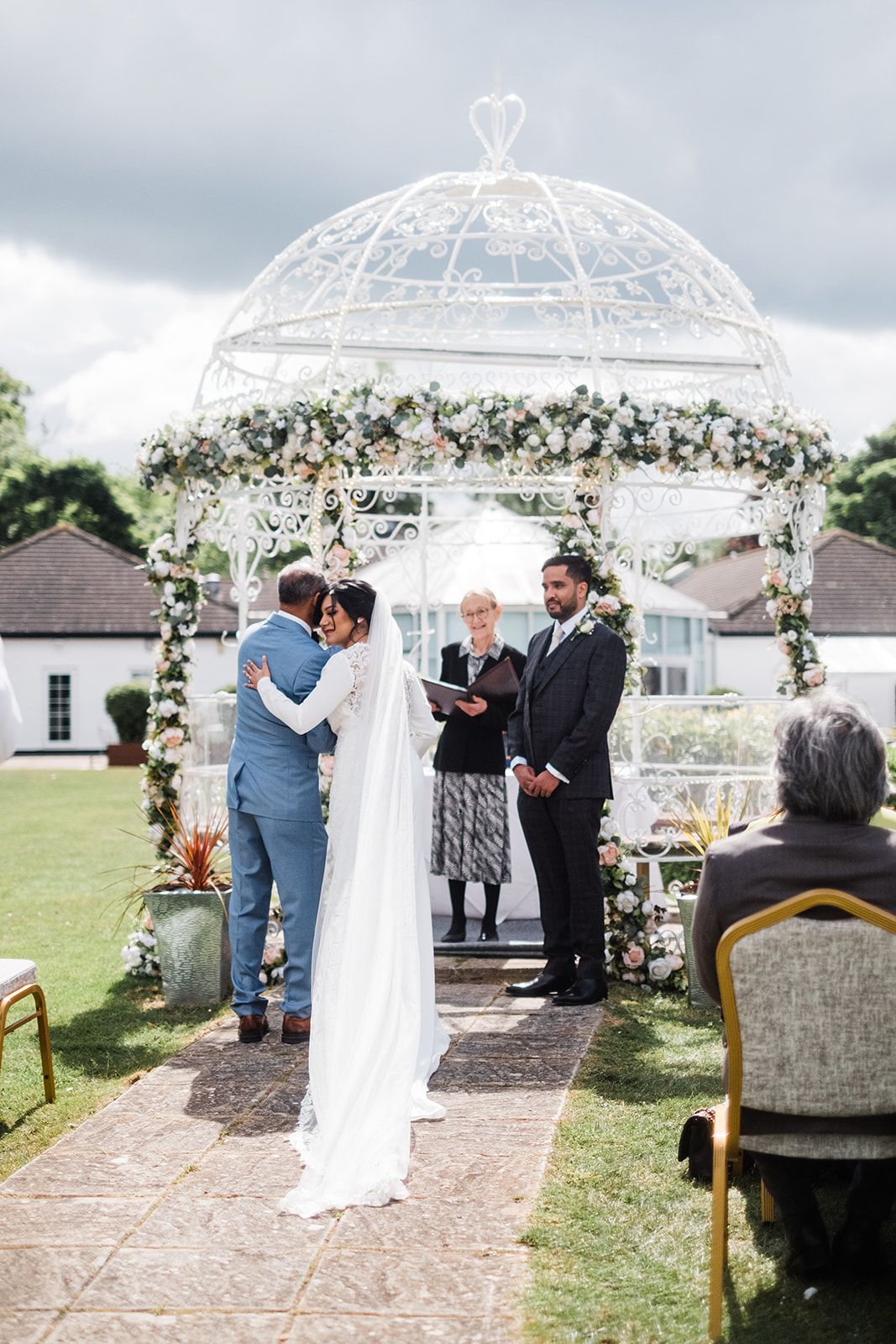 Couple sharing first kiss at outdoor wedding ceremony under white floral gazebo with officiant and guests.