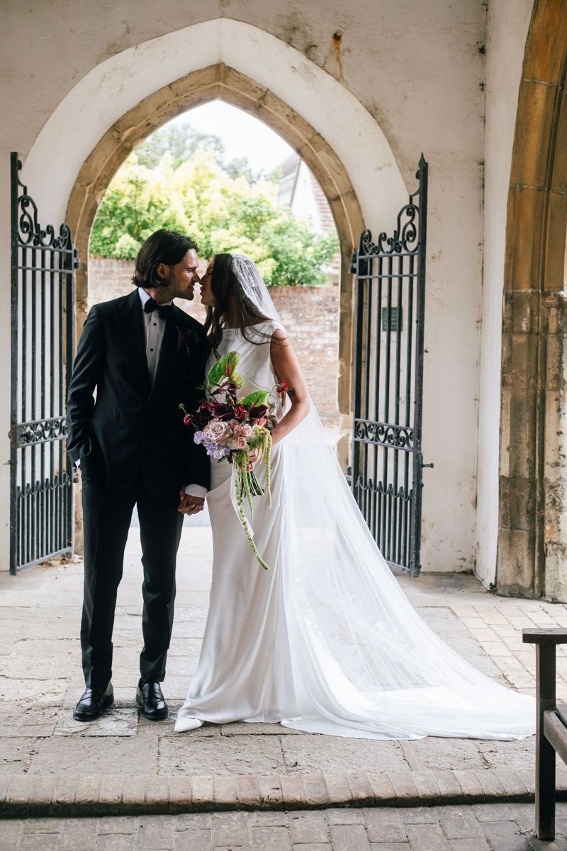 Newlyweds share romantic moment under stone Gothic arch at church entrance with bouquet.