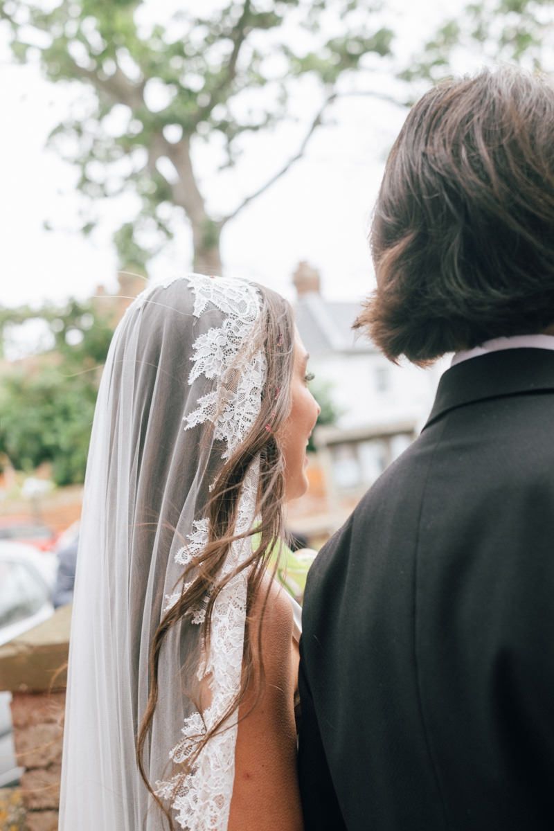 Bride in lace-trimmed veil gazes at groom outdoors after wedding ceremony.