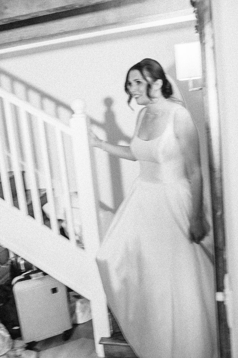 Bride in white wedding gown descending staircase, black and white film photography.
