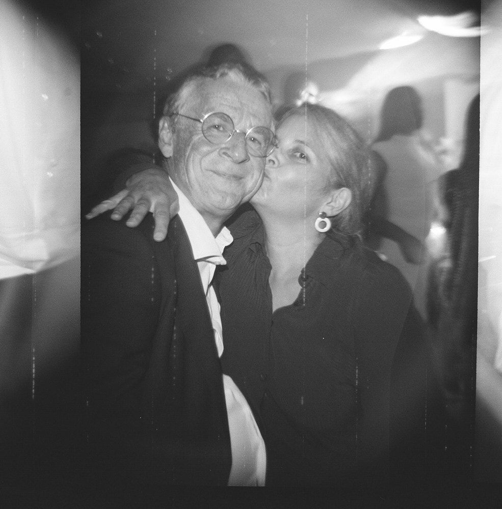Black and white photo of an older couple at a party, woman kissing mans cheek, both smiling warmly.