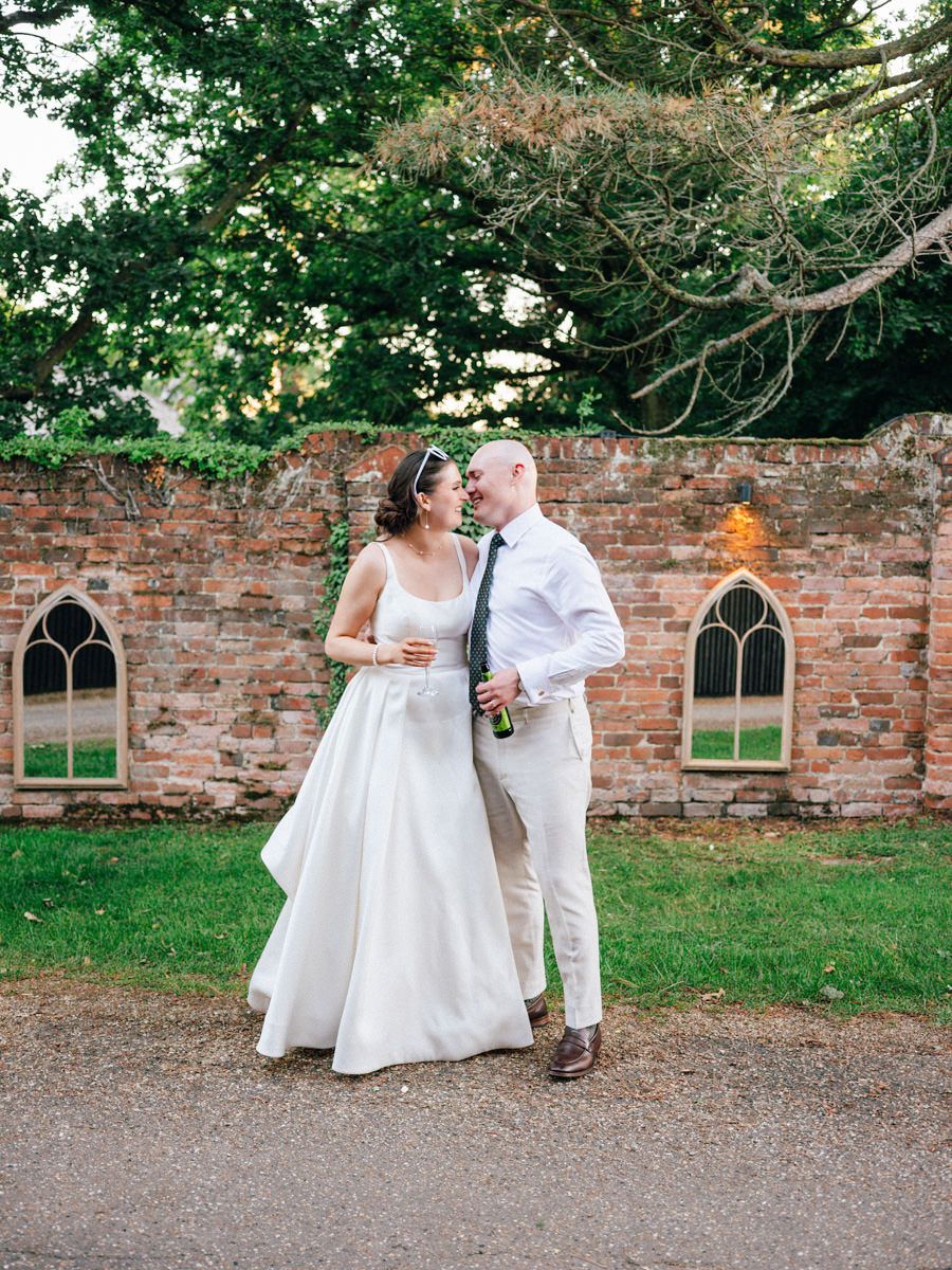 Bride and groom share a tender moment beside an ancient brick wall in a lush garden setting.