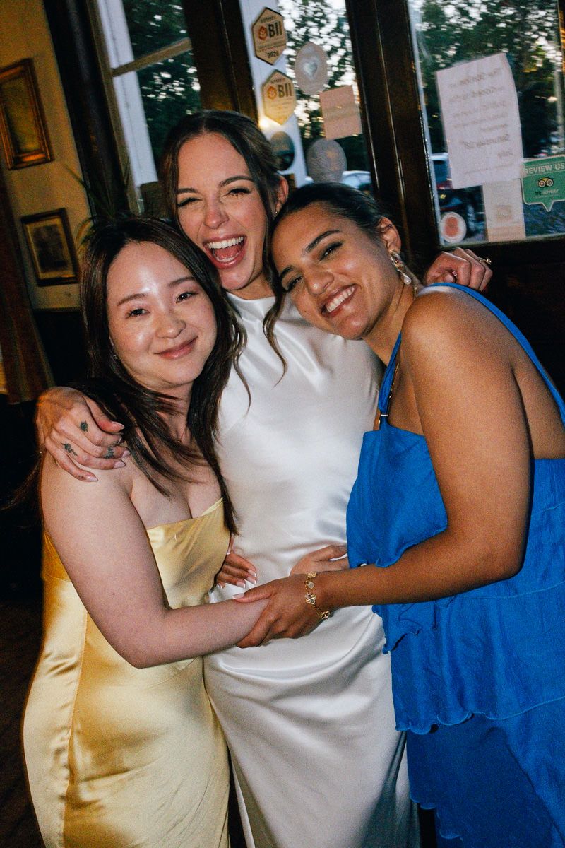A bride in a white dress laughs joyfully with two friends at her wedding reception.