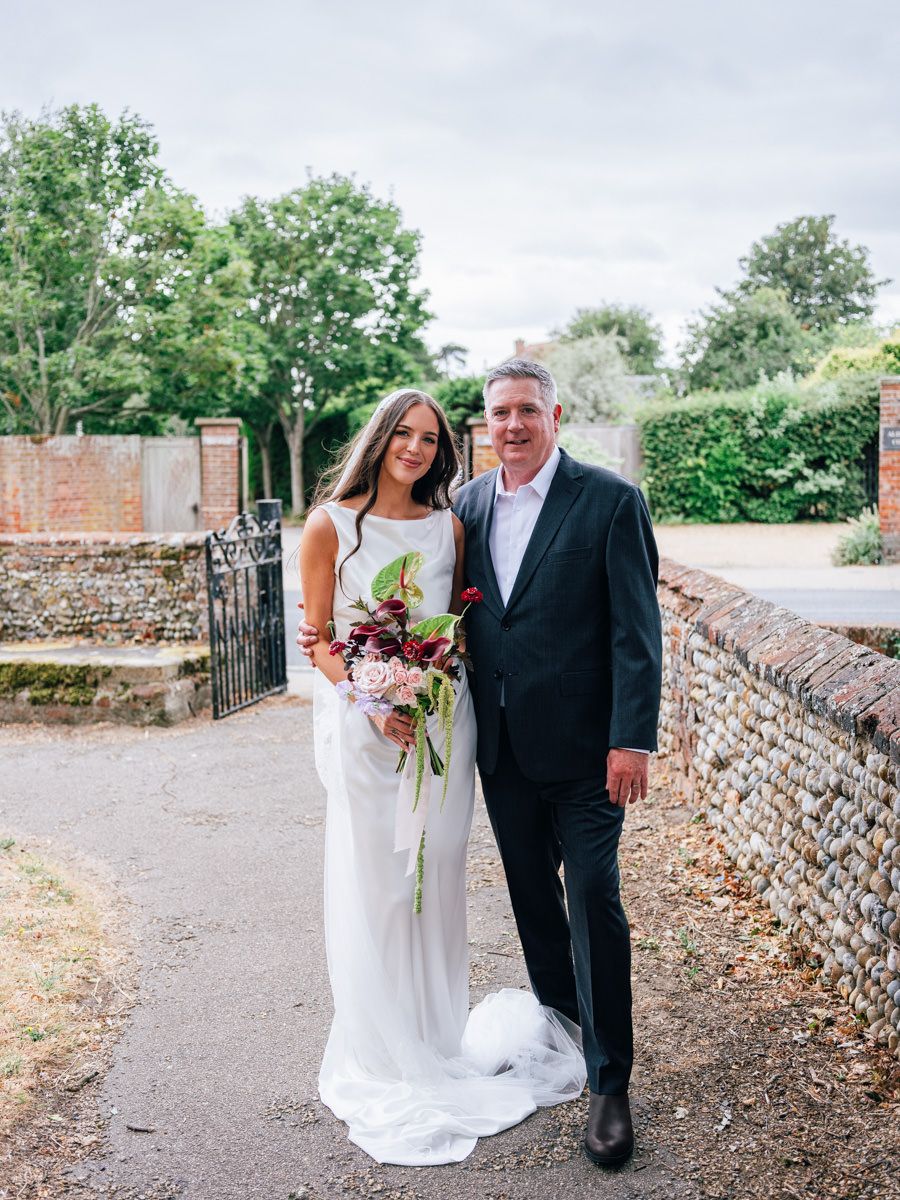 Bride in elegant white gown holding bouquet posing with father outside church.
