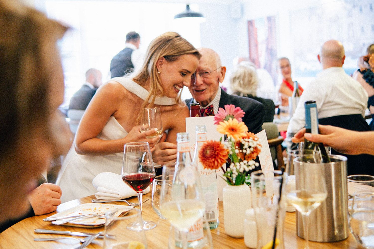 Bride in white dress sharing a joyful moment with elderly guest at wedding reception table with flowers and wine glasses.