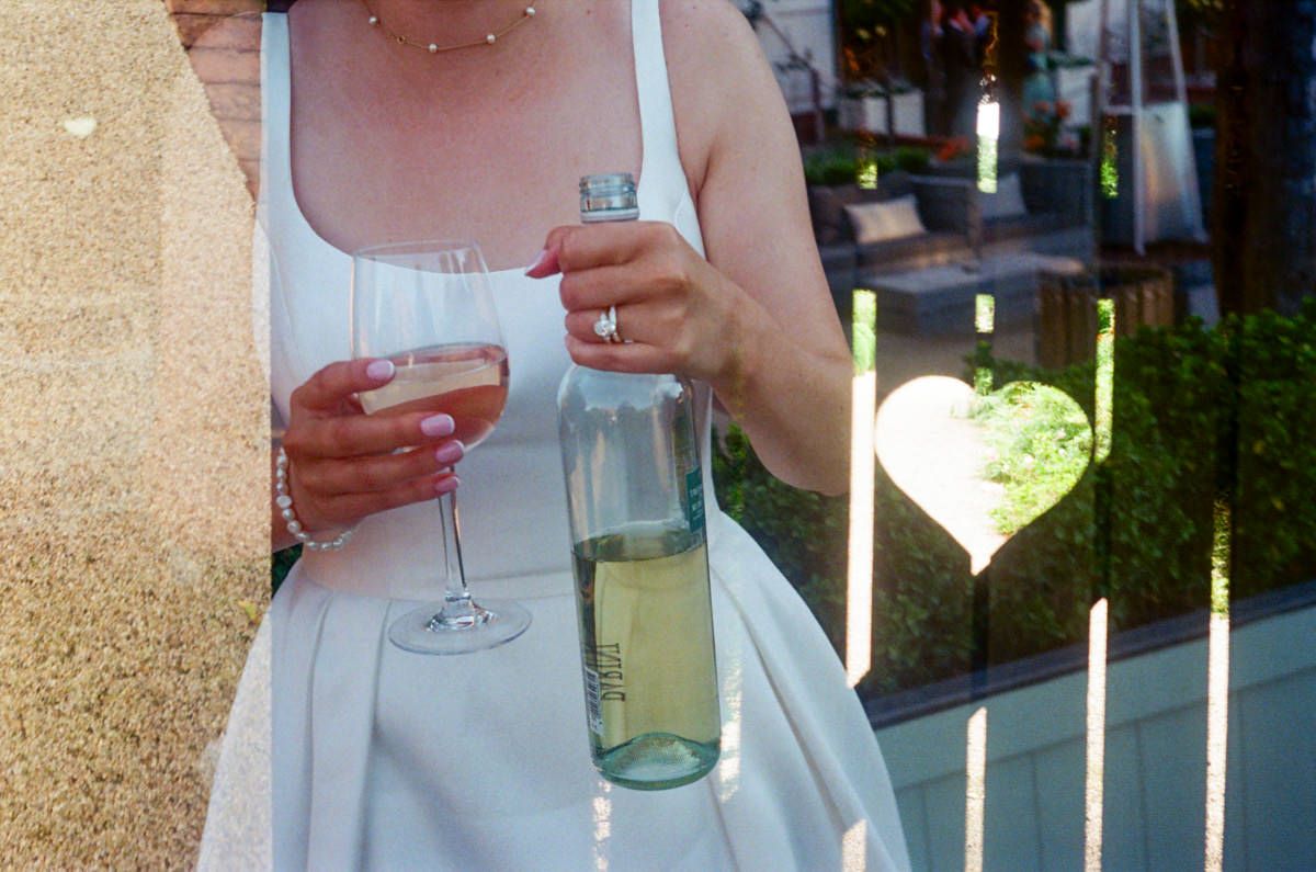 Bride holding a wine bottle and glass at an outdoor wedding reception with heart lights.