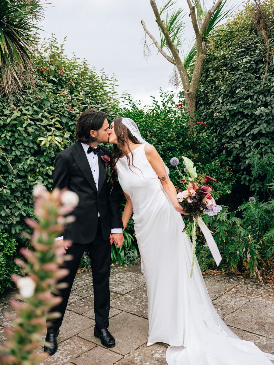 Bride and groom sharing a romantic kiss in a garden, bride holding a large floral bouquet.