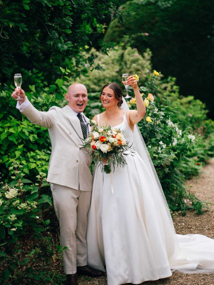 Newlyweds celebrating with champagne glasses raised in lush garden after wedding ceremony.