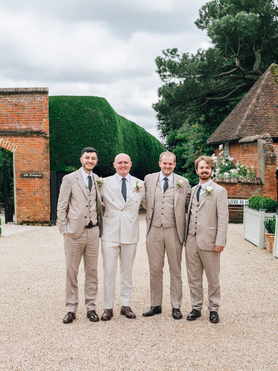 Groom and three groomsmen posing in tan suits outside a brick wedding venue.