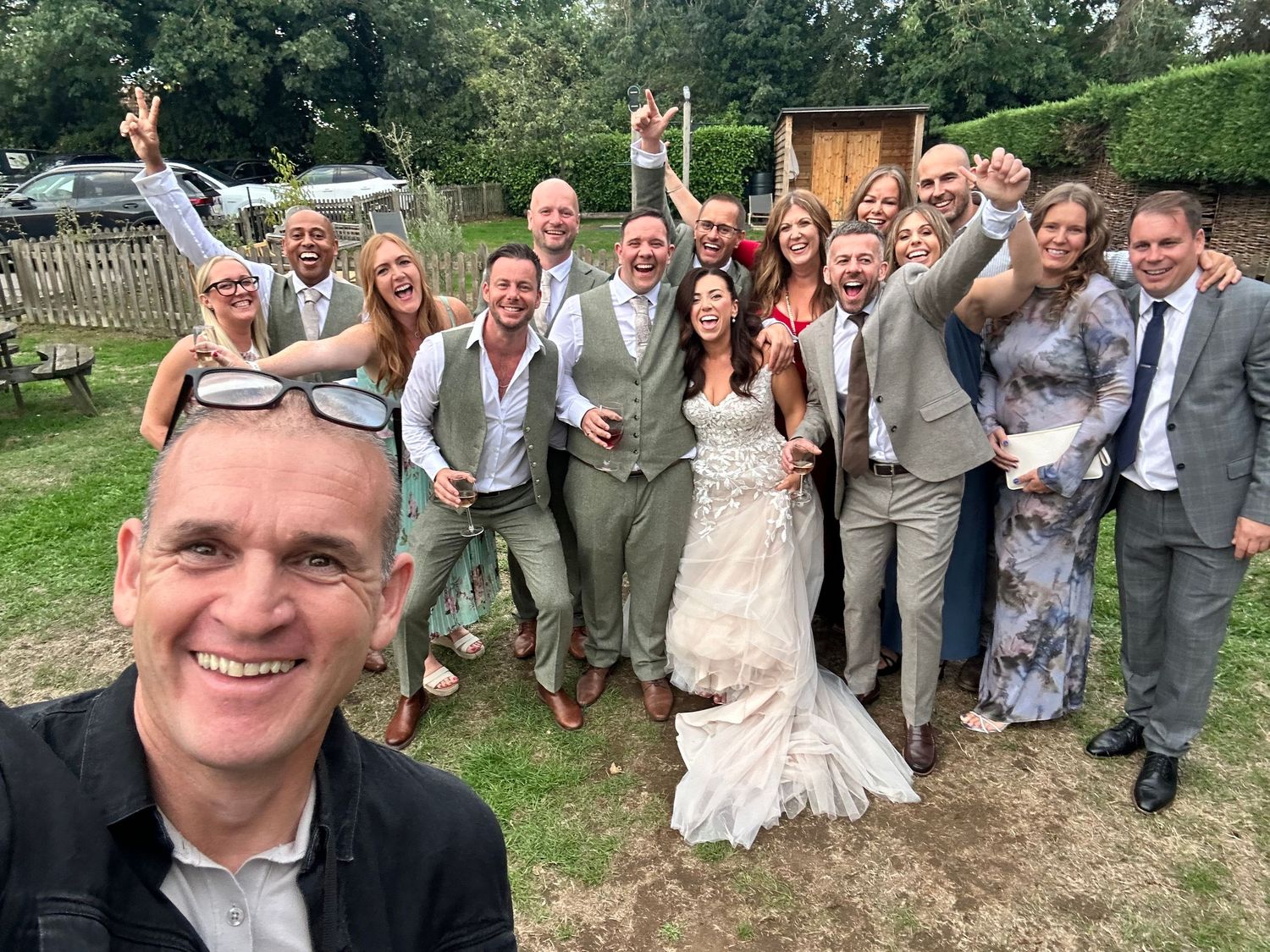 A joyful wedding group selfie outdoors with bride in white gown surrounded by guests in suits and dresses celebrating.