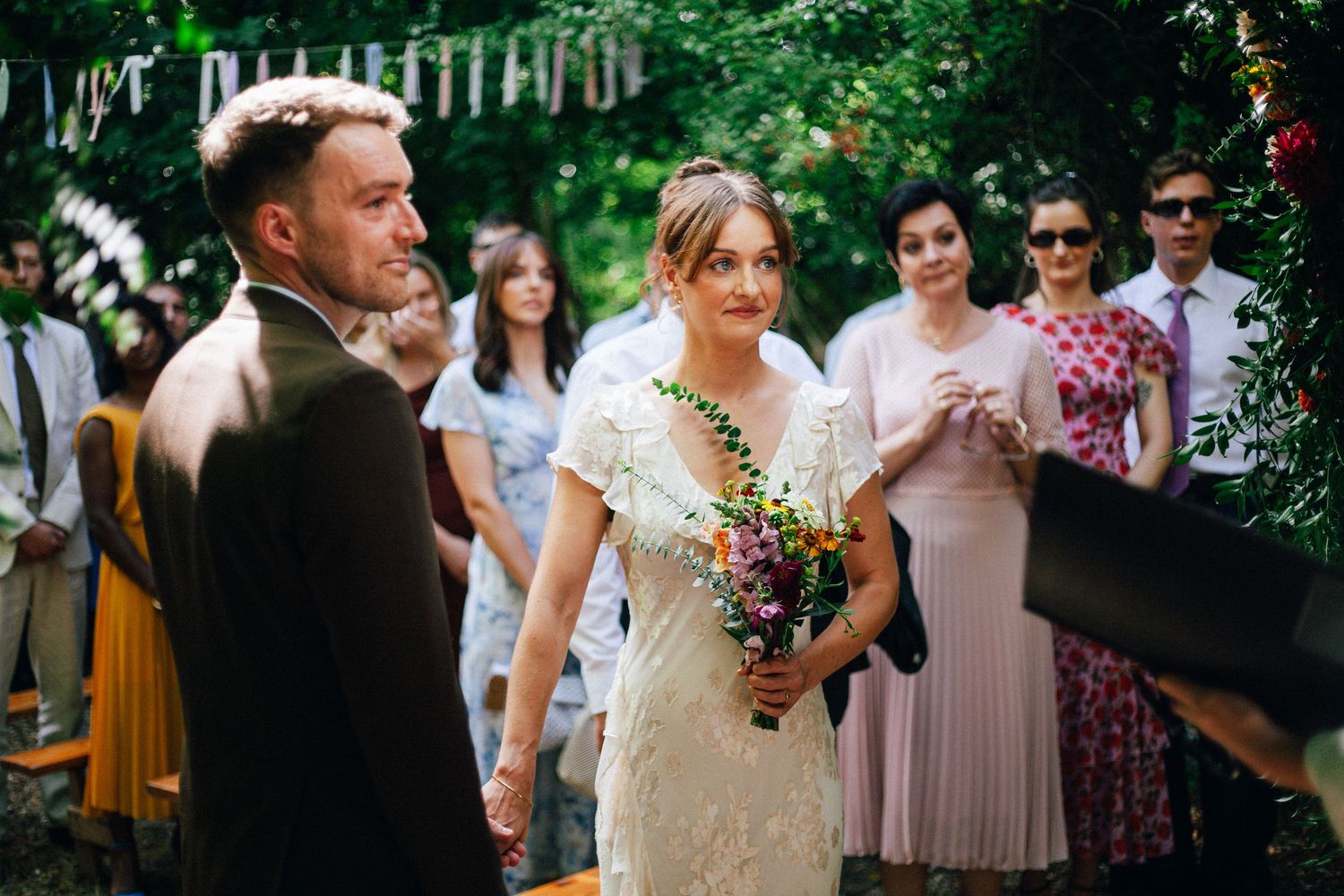 Bride in white lace gown holds colorful wildflower bouquet at outdoor garden wedding ceremony with guests.