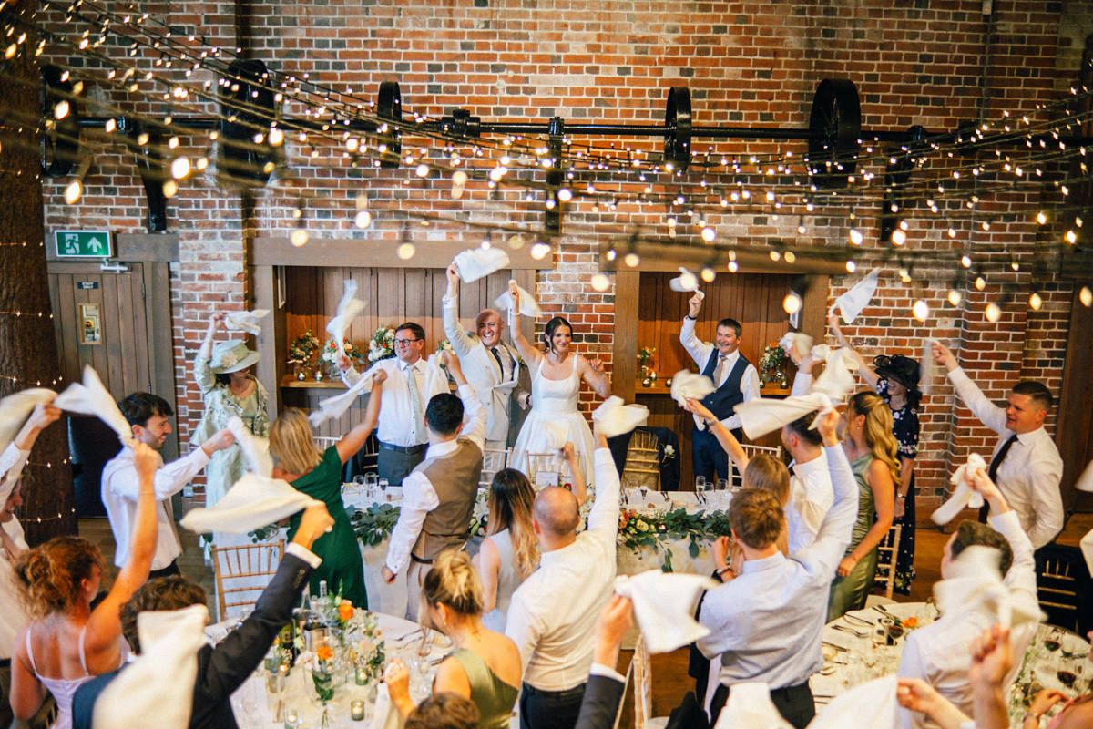 Joyful wedding reception guests celebrating with confetti under warm string lights in a rustic brick venue.