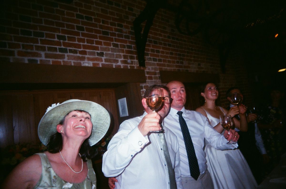 Wedding guests raise glasses in a celebratory toast inside a rustic brick-walled venue.