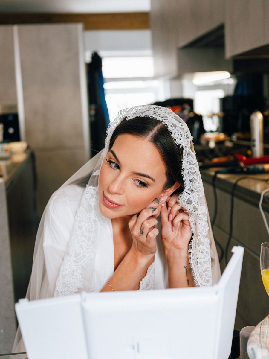 Bride in a white robe putting on earrings while wearing a lace mantilla veil before the wedding.