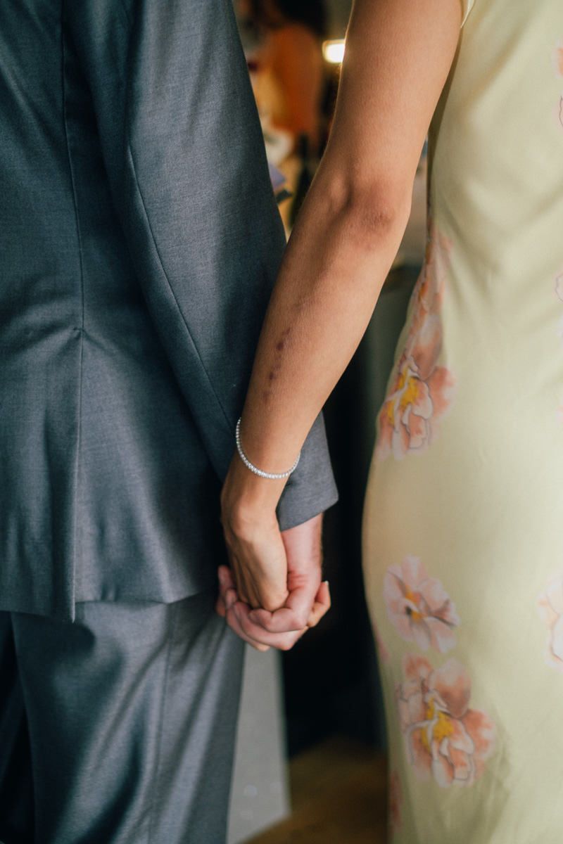 Close-up of bride and groom holding hands, bride wearing pearl bracelet in floral dress at wedding.