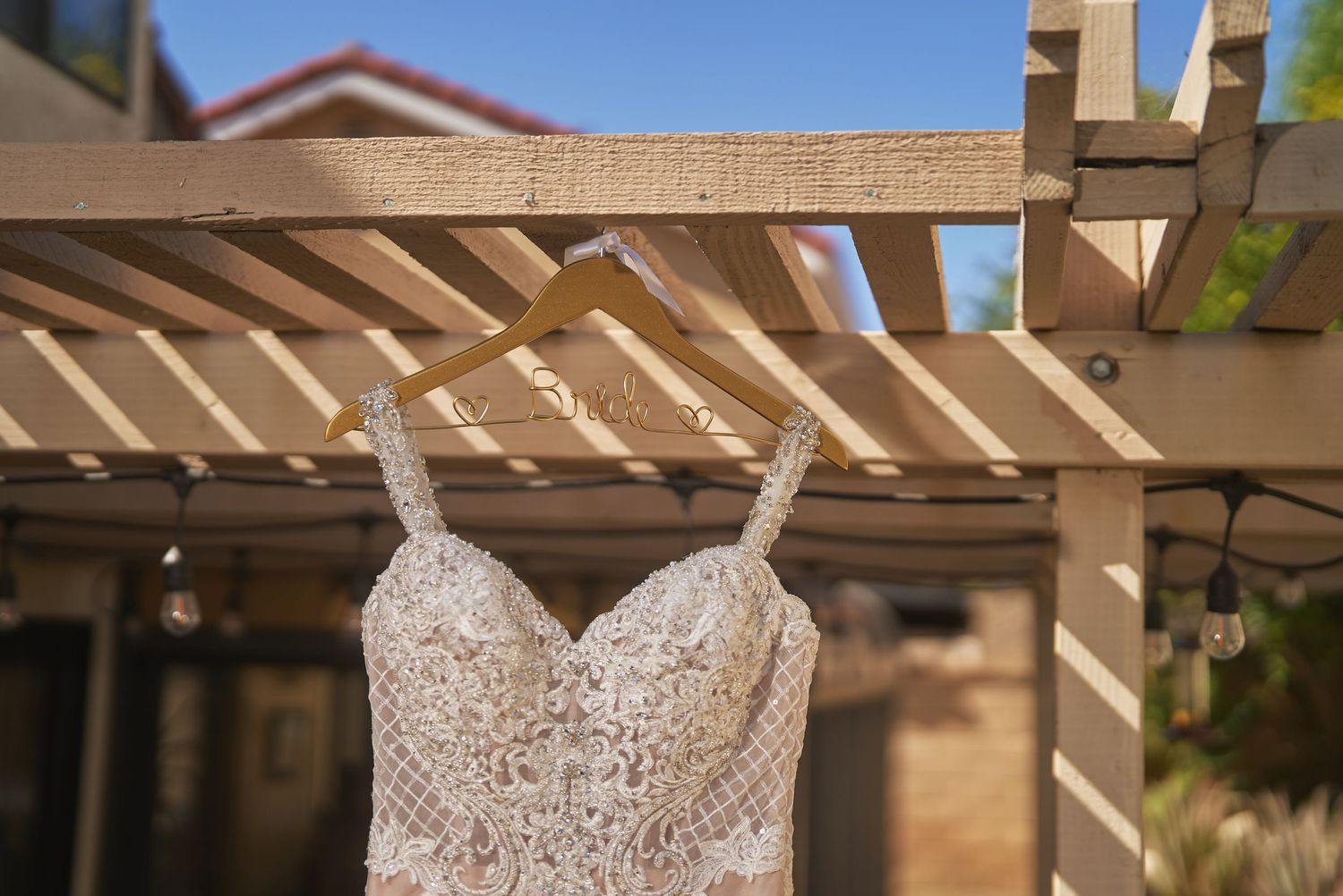 Elegant lace wedding dress hanging on a wooden pergola under a clear blue sky.