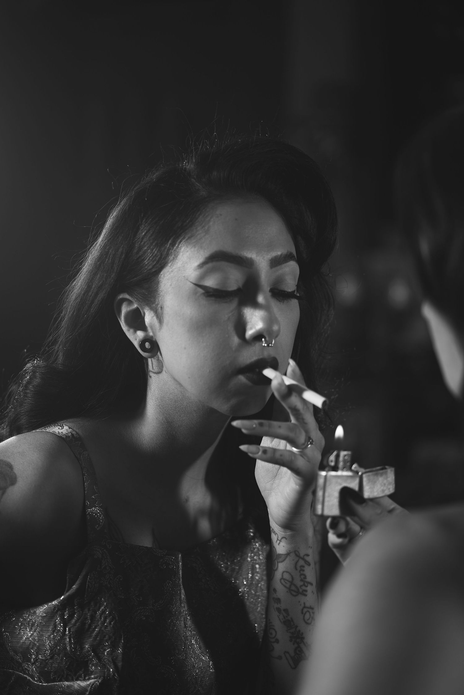 Black and white portrait of woman with long hair lighting cigarette in evening wear.