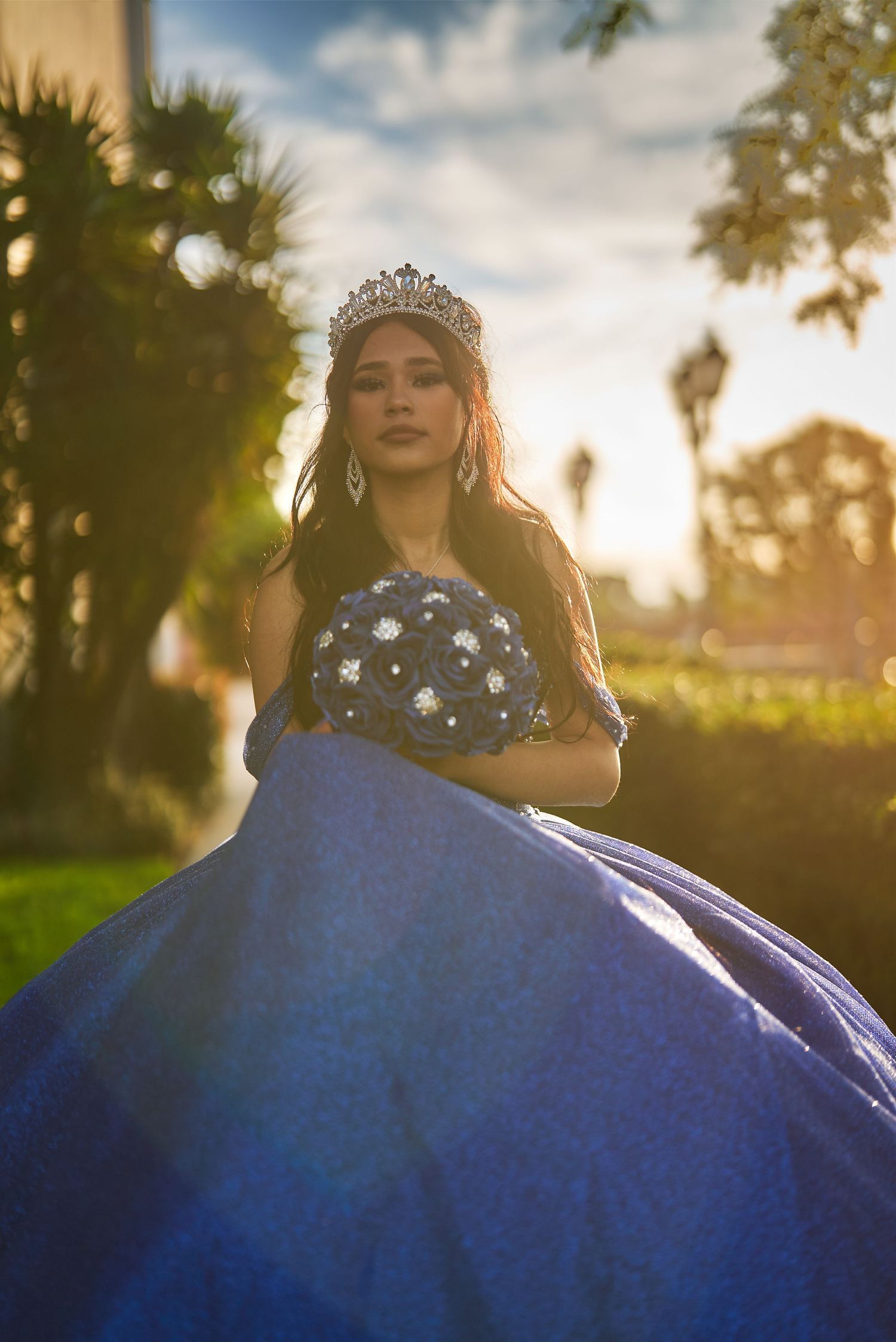 Woman in royal blue ball gown and tiara poses against sunset sky with palm trees.
