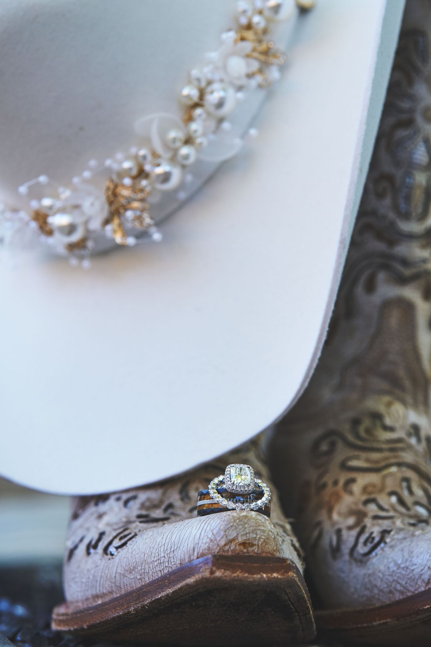 Diamond engagement ring resting on cowboy boots beside a white hat adorned with pearls and gold floral accents.