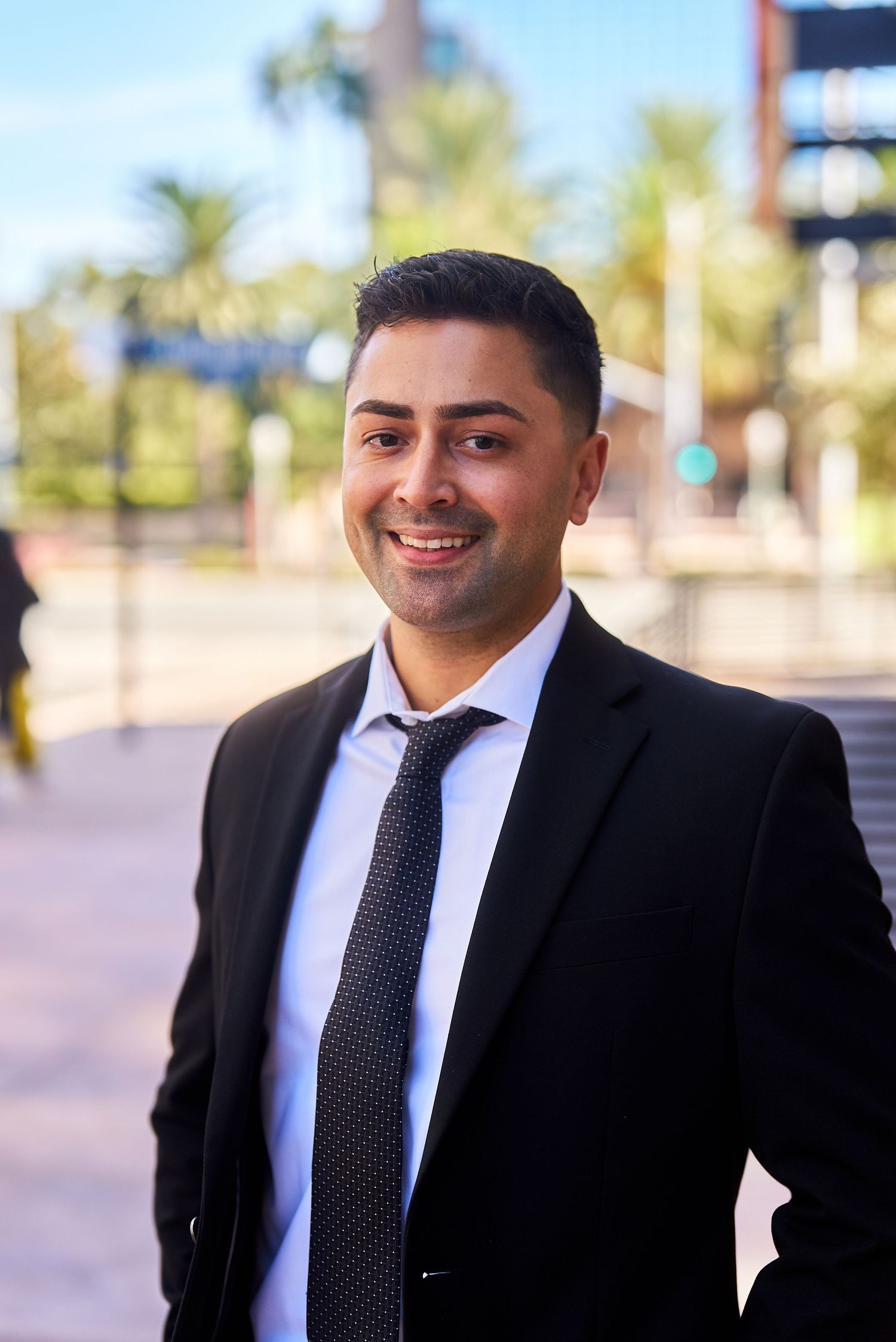 Professional in dark suit and tie smiling against urban background with palm trees.