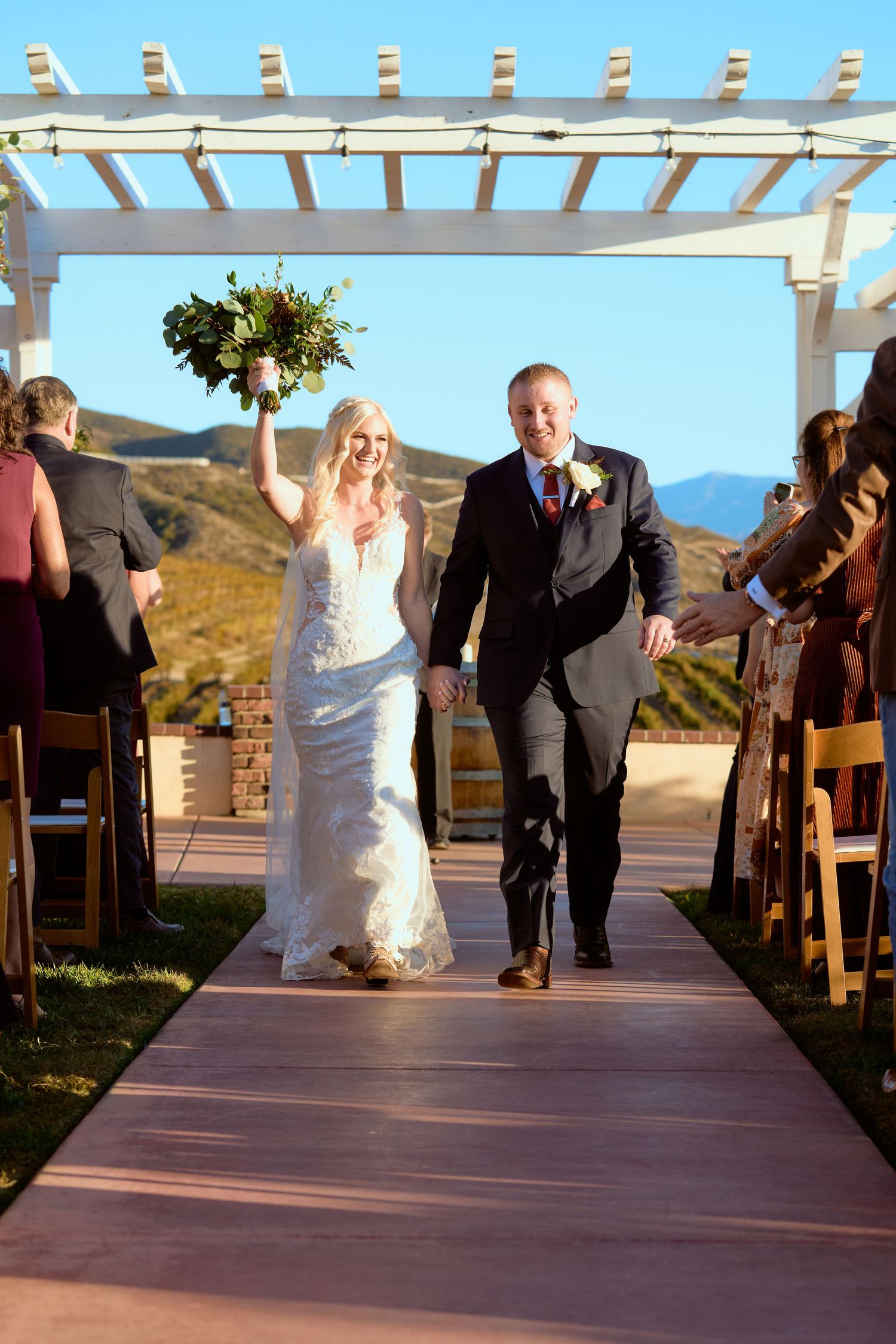Joyful newlyweds walk down outdoor aisle, bride raising bouquet, mountains backdrop, white pergola, guests cheering.