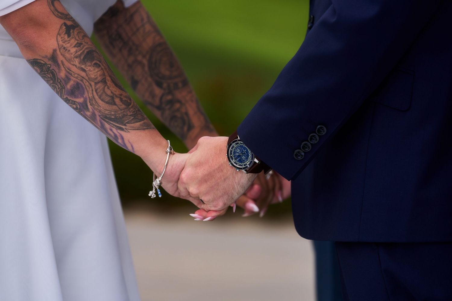 Couple holding hands at wedding, bride with tattooed arm wearing bracelet, groom in navy suit with blue watch.