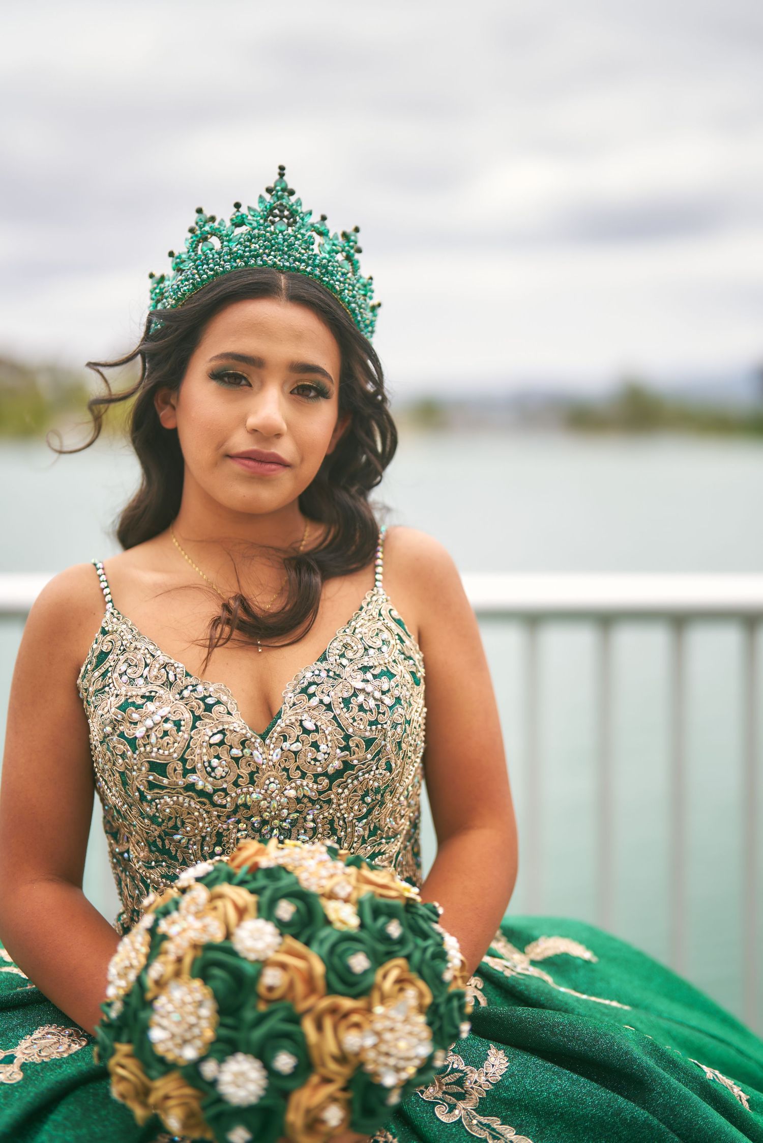 Woman wearing a green quinceañera dress and ornate turquoise crown poses outdoors by a waterfront railing.