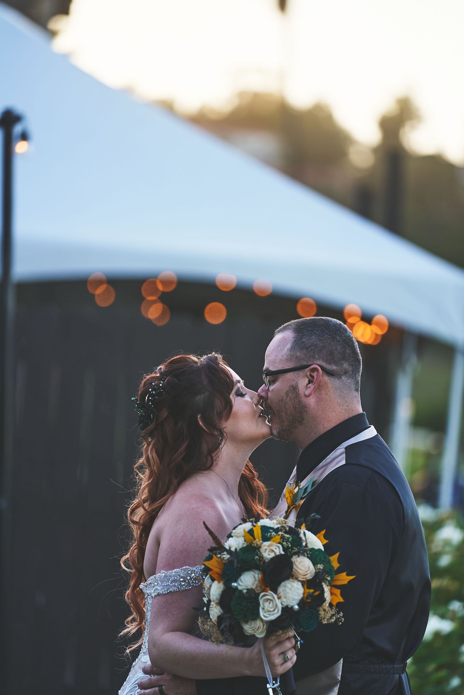 Wedding couple sharing a romantic kiss at sunset with bouquet of white flowers.