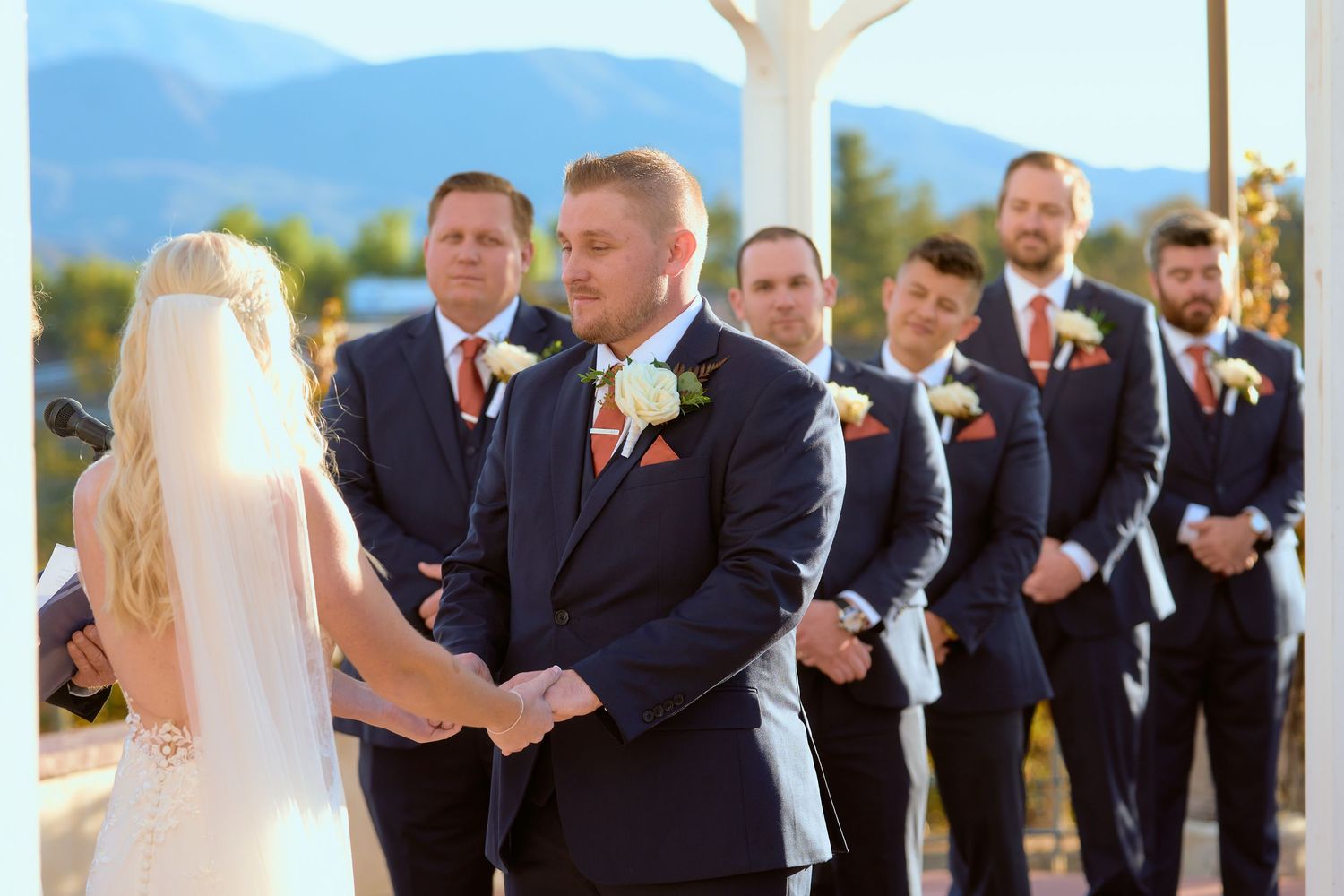 Groom in navy suit holds hands with bride at outdoor wedding ceremony with groomsmen and mountains in background.