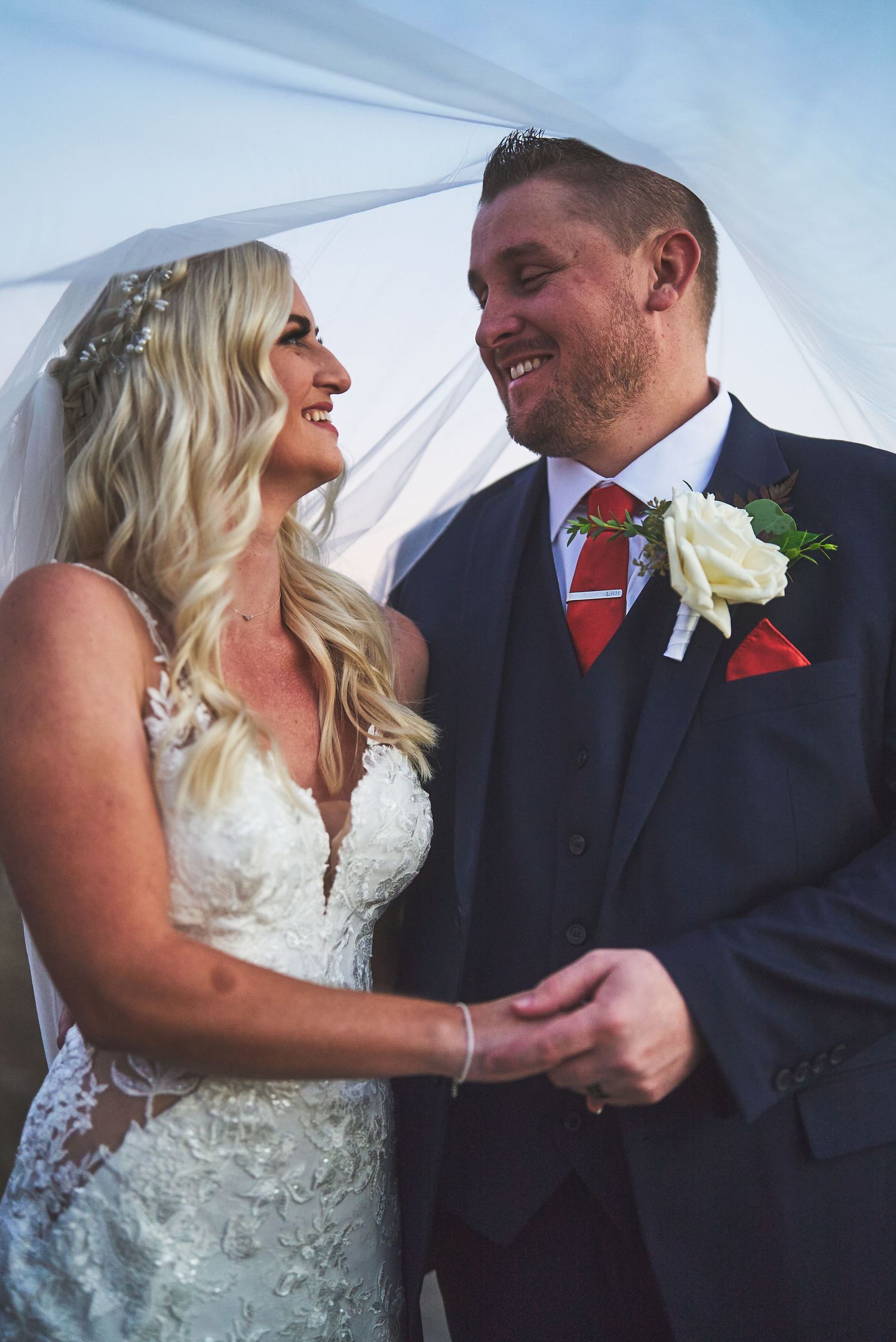 Bride and groom smiling at each other under a veil, wearing a lace wedding dress and navy suit with red tie.