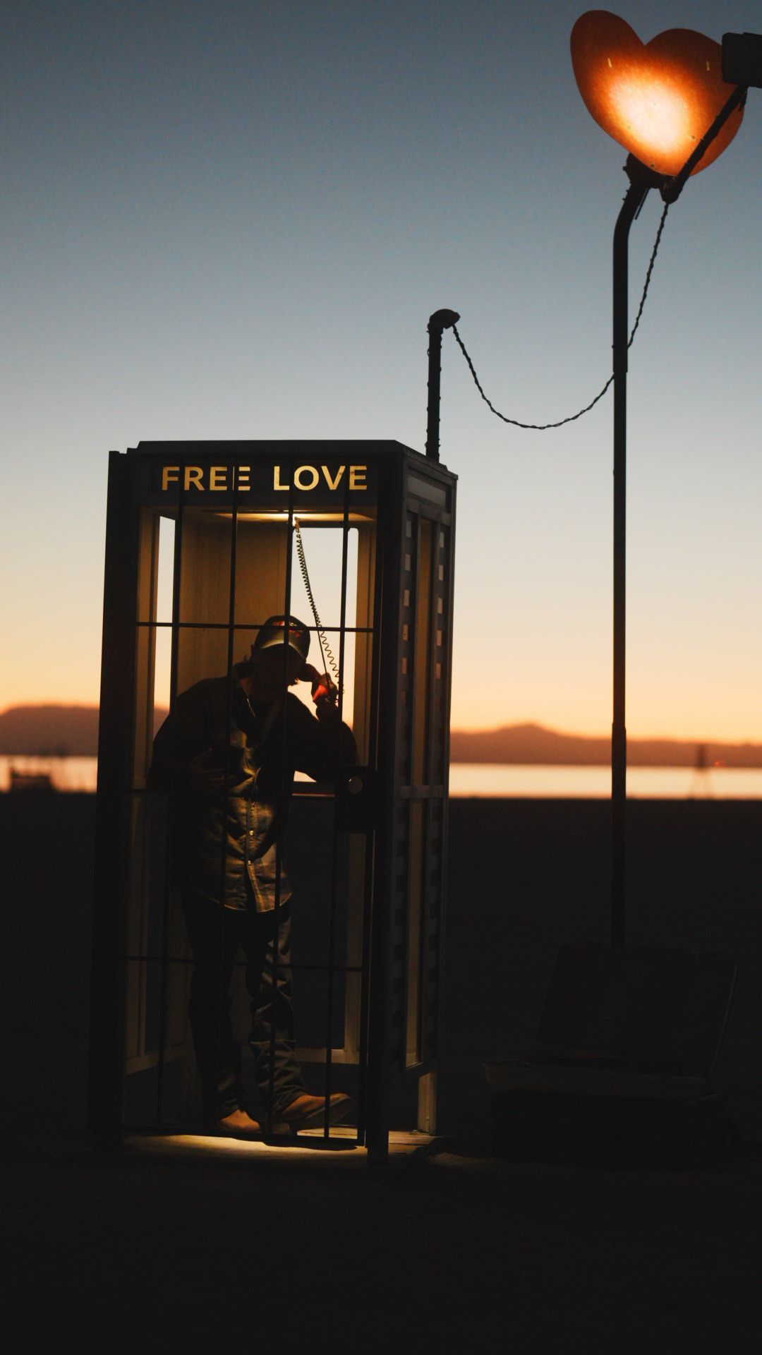 Silhouette in phone booth with heart-shaped light and 'FREE LOVE' text at sunset.