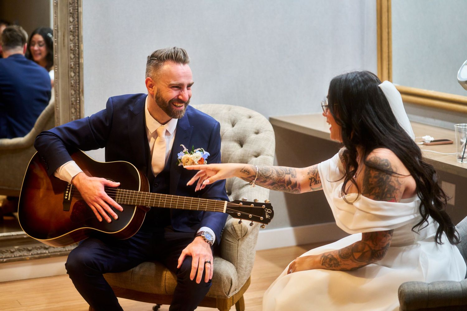 Groom in navy suit plays acoustic guitar for tattooed bride in white dress during intimate wedding reception moment.