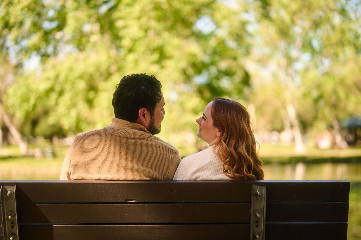 Couple sitting together on a park bench, facing each other surrounded by lush green trees near a pond.