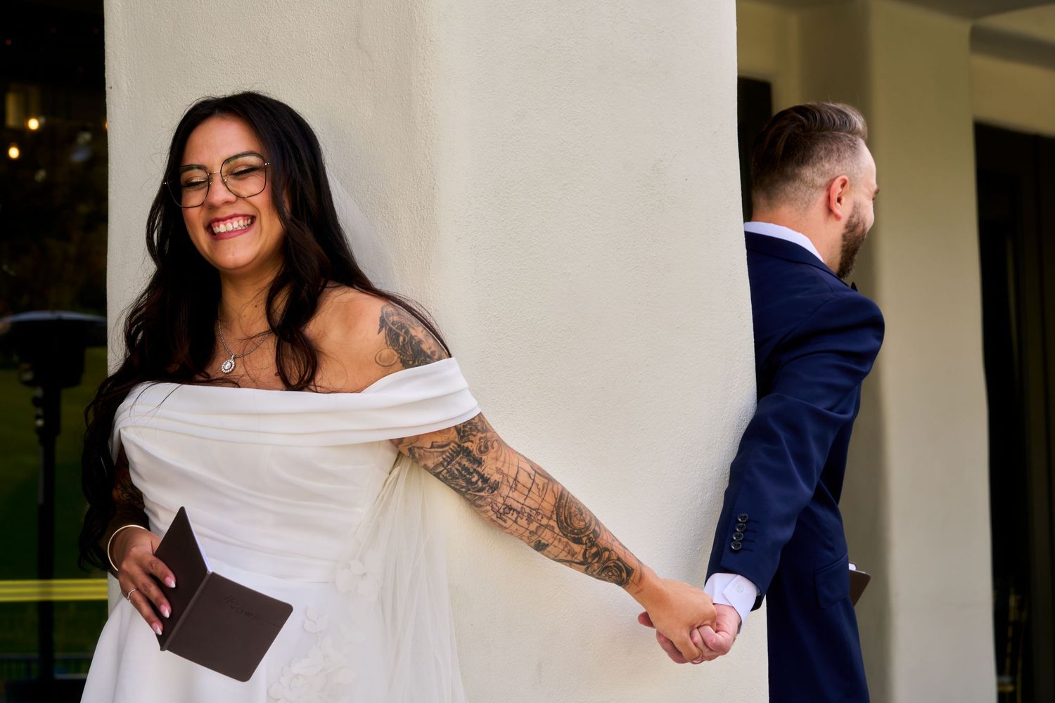 Bride in white off-shoulder gown with tattoos holds groom's hand around a pillar during a first look wedding moment.