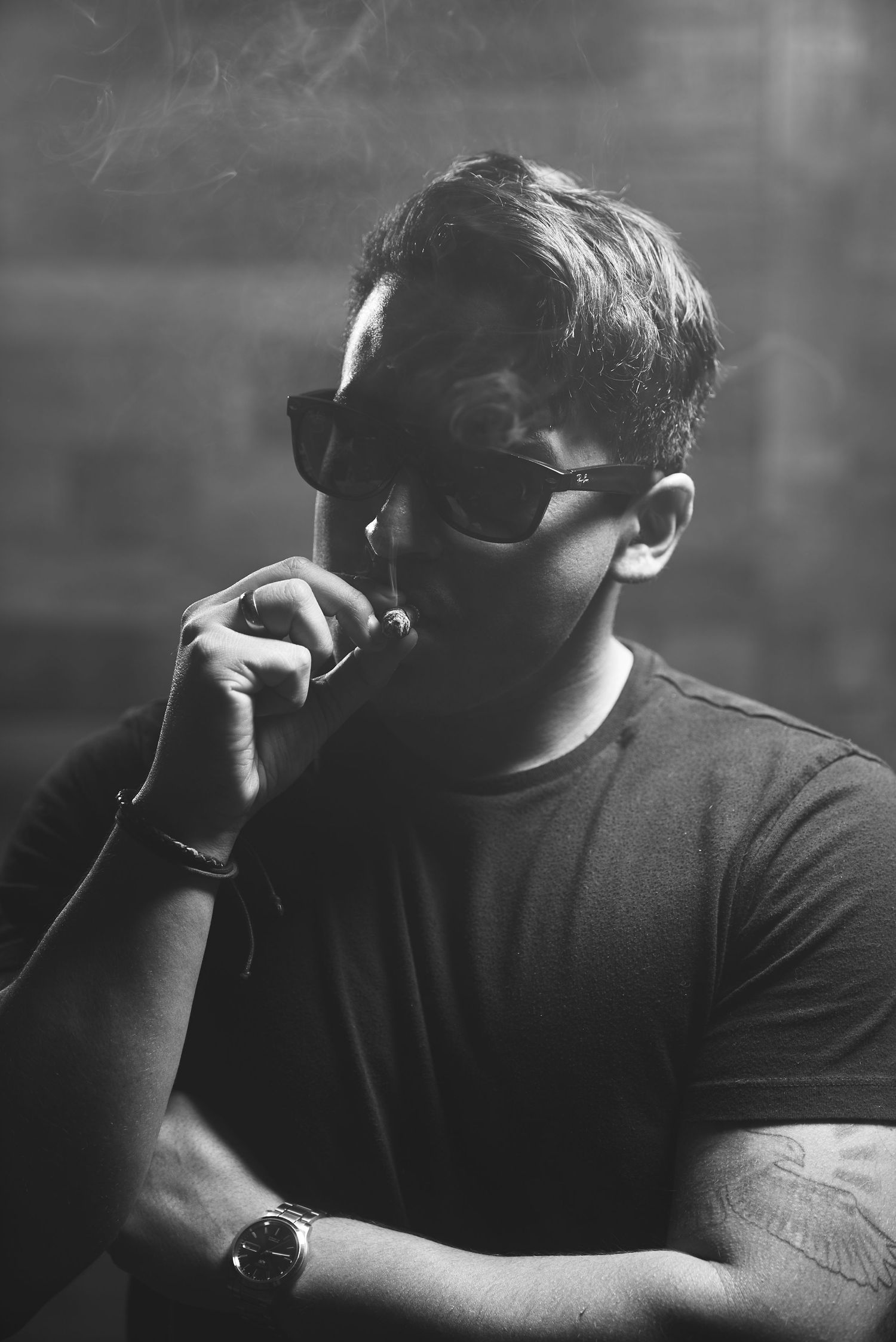 Dramatic black and white photo of person in sunglasses smoking while wearing casual attire.