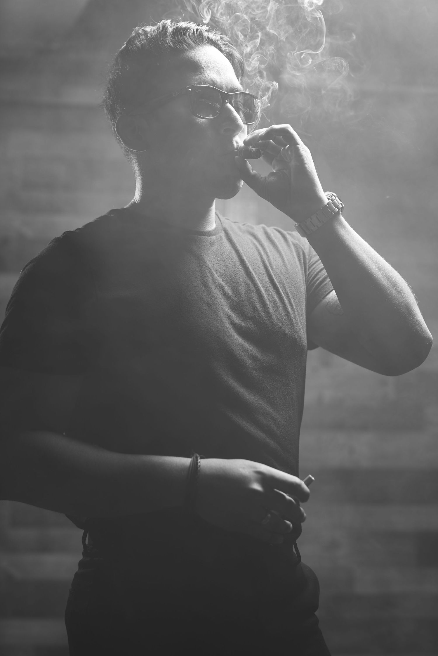 Artistic black and white photo of person smoking in dramatic lighting against dark wall.