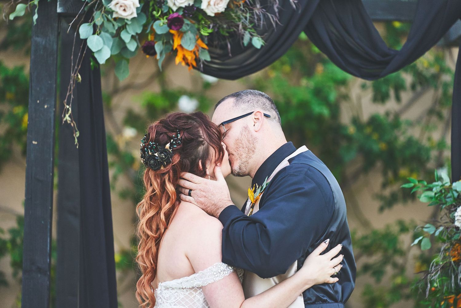 Bride and groom share first kiss under floral arch with black draping at outdoor wedding ceremony.