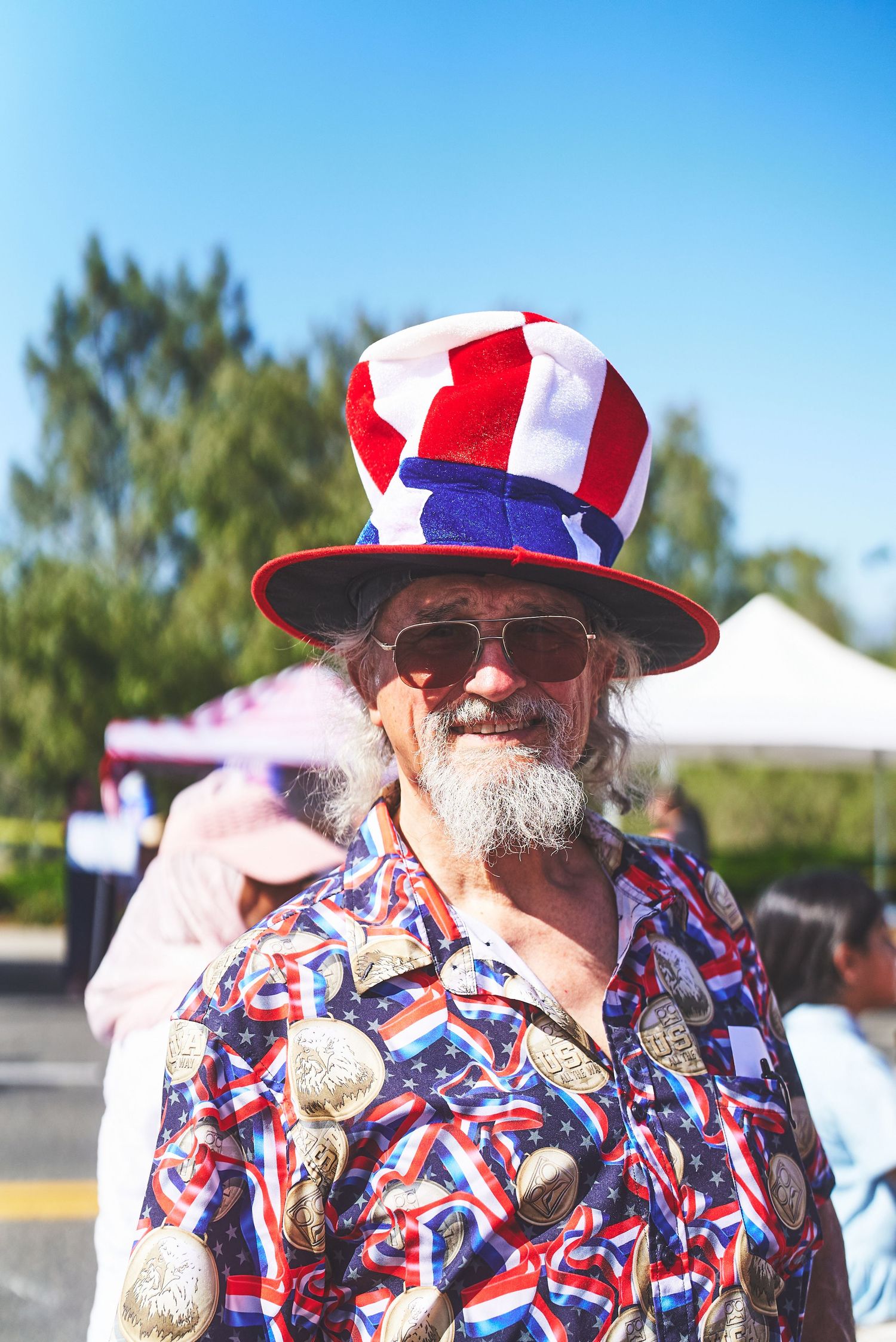 Man wearing patriotic Uncle Sam hat and flag-print shirt at outdoor event.