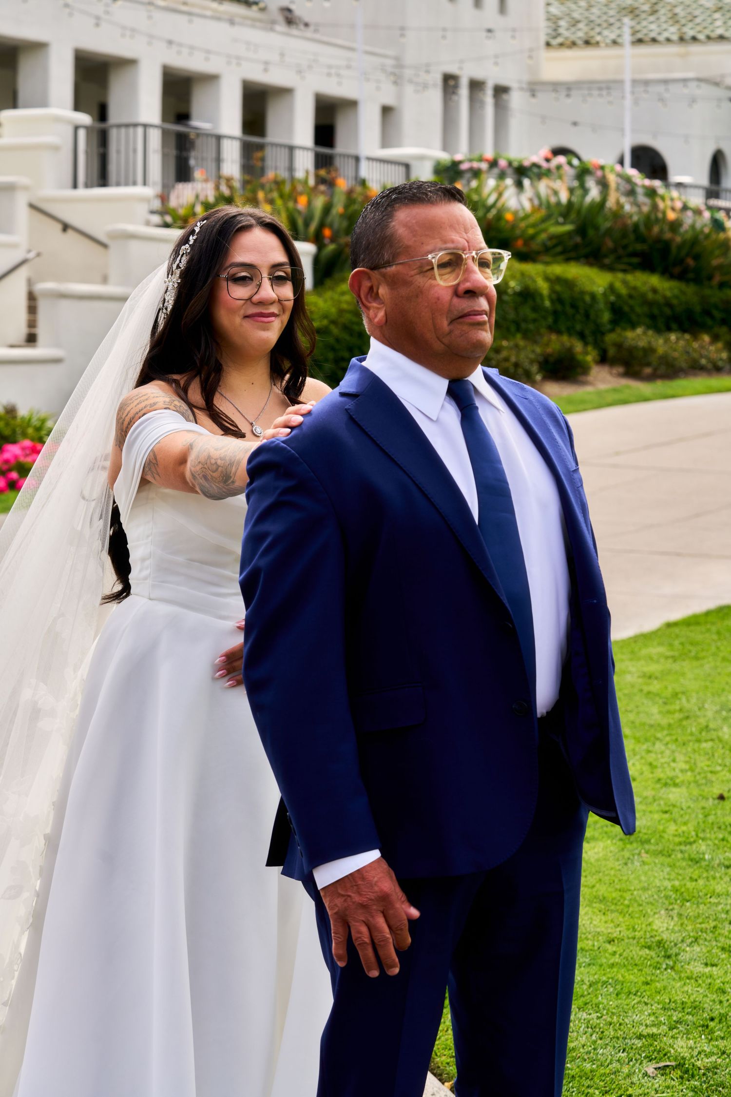 Bride in white gown and veil with father in blue suit walking together outside a elegant venue.