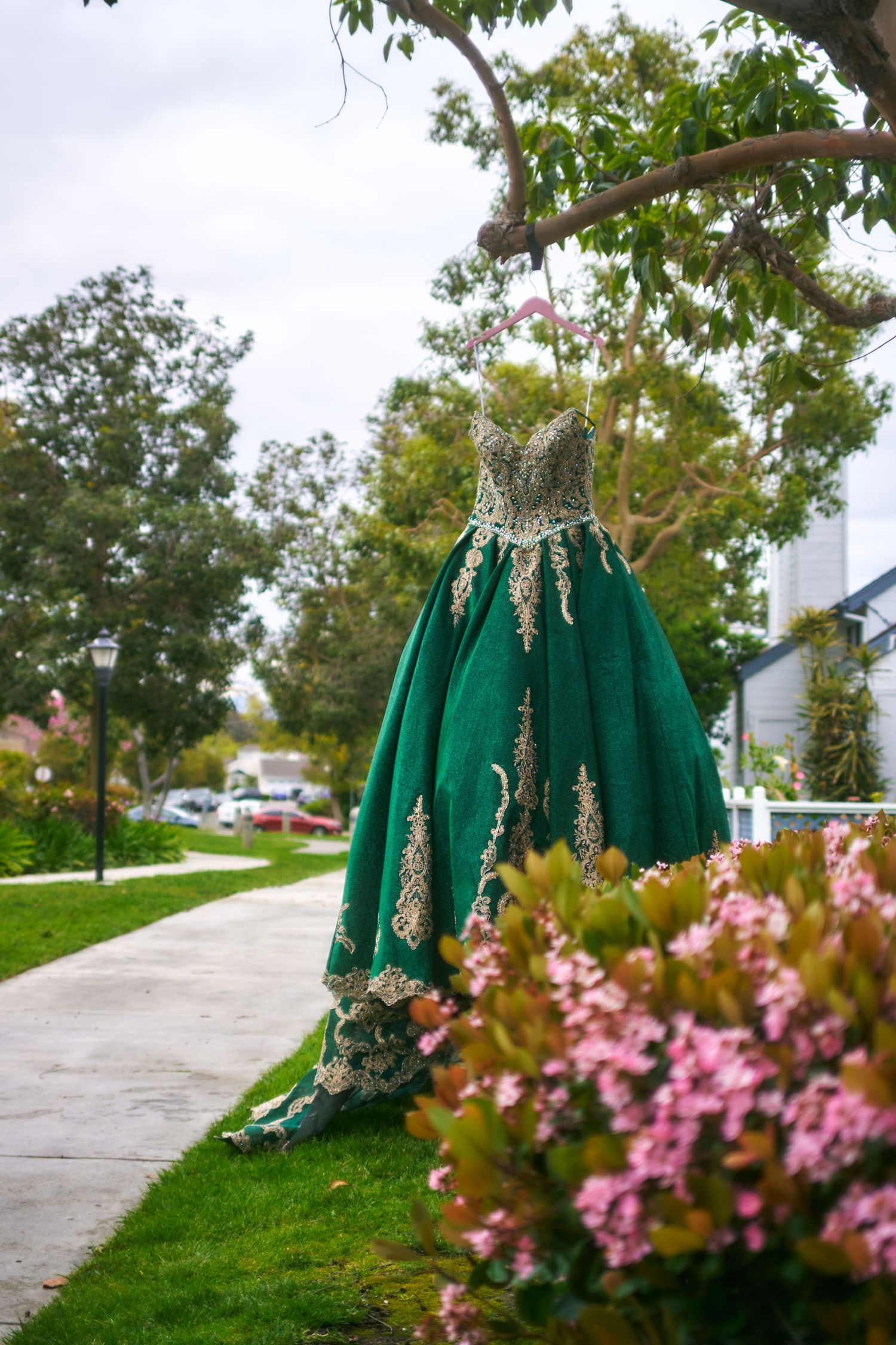 Elegant emerald green ball gown with gold embellishments displayed outdoors next to pink flowers on a sidewalk.